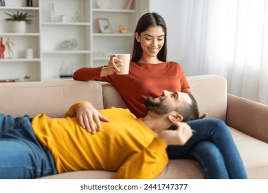 A cheerful young woman holds a coffee mug while a man lies with his head on her lap, both enjoying a cozy moment on a sofa