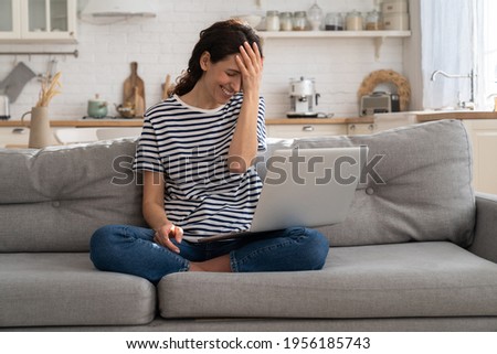 Cheerful young woman laugh while watching funny video on laptop at home sitting with crossed legs on sofa. Overjoyed happy female excited of online call or chat, astonished with good positive news