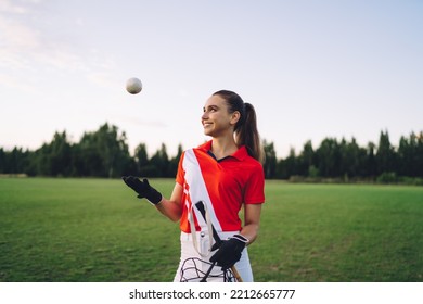 Cheerful Young Woman With Long Hair In Ponytail Wearing Red T Shirt And Gloves Standing On Green Meadow And Throwing Polo Ball Up In Air Against Forest On Sunset