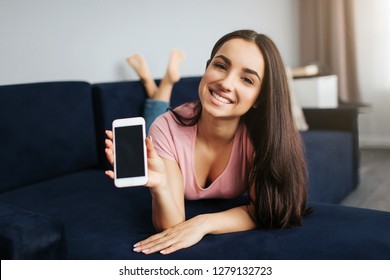 Cheerful young woman lying on stomach on sofa. She hold white phone and smiling to camera. Model show device with dark screen.