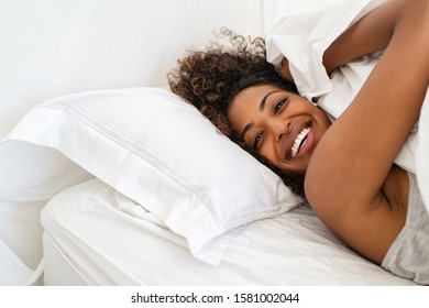 Cheerful young woman lying on bed playing with blanket and looking at camera. African girl feeling fresh after nap on bed with copy space. Laughing woman having fun while embracing pillow and blanket.