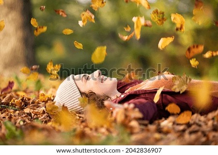 Cheerful young woman lying over leaves with closed eyes and smiling. Woman lying over autumn ground relaxing at park. Casual joyful girl having fun throwing leaves in autumn park with copy space.