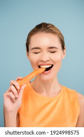 Cheerful Young Woman With Natural Makeup Biting Whole Carrot Isolated On Blue