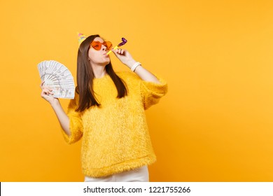 Cheerful Young Woman In Orange Heart Glasses, Birthday Hat With Playing Pipe Holding Bundle Lots Of Dollars Cash Money, Celebrating Isolated On Yellow Background. People Sincere Emotions, Lifestyle