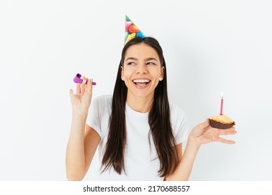 Cheerful Young Woman In Party Cap Celebrating Birthday Holding Kazoo And Cake With Candle In Her Hands, White Background