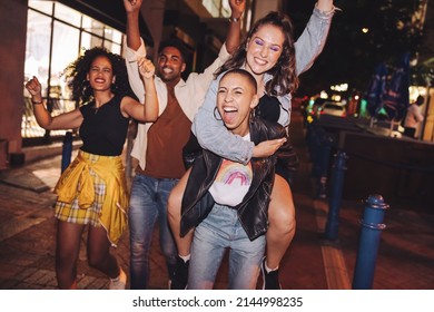 Cheerful young woman piggybacking her friend in the city. Girlfriends cheering and having fun while going out with their friends at night. Group of vibrant friends hanging out together on the weekend.