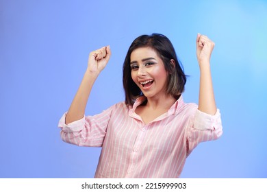 Cheerful young woman in pink shirt raising hands up   smiling excited  triumphing over winning prize  celebrating victory  standing blue gradient background