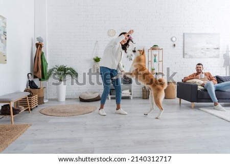 cheerful young woman playing with akita inu dog near bearded boyfriend with cup of coffee sitting on couch