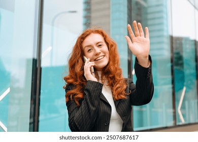 Cheerful young woman with red hair talking on her phone and waving outside modern building during daylight - Powered by Shutterstock