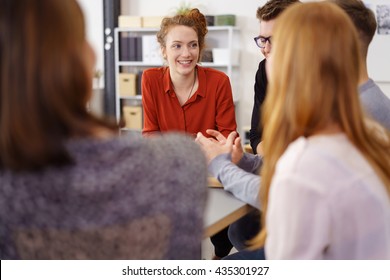 Cheerful Young Woman In Red Shirt Listening To Coworkers Discuss Something During Meeting Around Large Table In Small Office