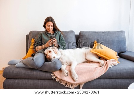 Cheerful young woman relaxing and enjoying leisure time at home, typing a text message using smart phone with her pet dog lying in her lap
