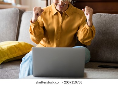 Cheerful Young Woman Showing Fist With Both Hands While Working On Laptop Sitting On Sofa At Home. Excited Woman Using Laptop While Woking. Happy Woman Relaxing On Sofa With Laptop On Table.