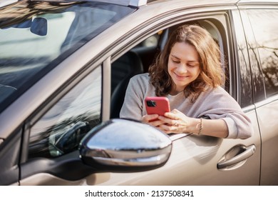 Cheerful young woman sitting in a car in the driver's seat looking into a smartphone, paying for parking and navigating in the city - Shutterstock ID 2137508341