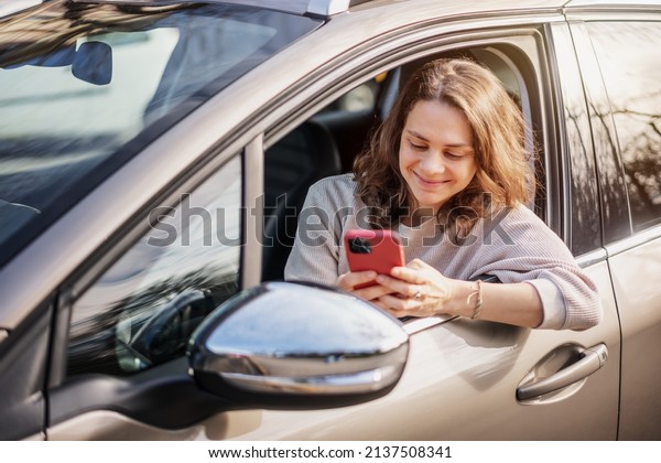 Cheerful young woman sitting in a car in the\
driver\'s seat looking into a smartphone, paying for parking and\
navigating in the city