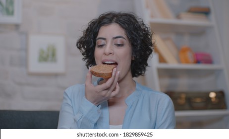 A Cheerful Young Woman Is Sitting At Home On An Armchair And Eating Peanut Butter Bread.