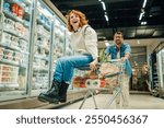 Cheerful young woman is sitting in a shopping cart while her boyfriend pushes her through the supermarket aisles, enjoying a playful moment together as they shop for groceries