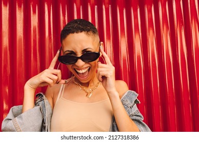 Cheerful young woman smiling at the camera while touching her sunglasses. Happy young woman standing against a red background. Young woman feeling vibrant and full of life. - Shutterstock ID 2127318386