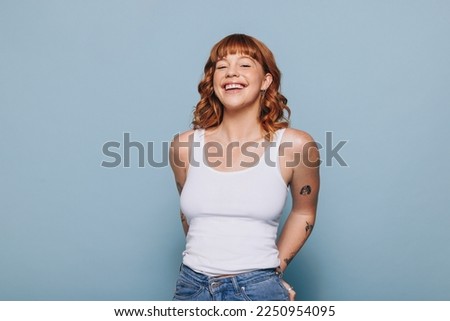 Cheerful young woman smiling at the camera while wearing a tank top and jeans in a studio. Portrait of a happy woman with ginger hair and tattoos standing against a blue background.