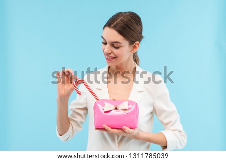 Cheerful young woman standing isolated over blue background, holding cosmetic bag, taking candy cane out