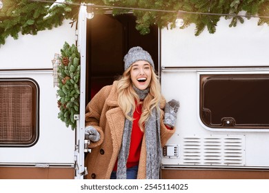 A cheerful young woman stands at the door of a festive camper, wearing winter attire. The entrance is adorned with greenery, showcasing a cozy, holiday spirit. - Powered by Shutterstock