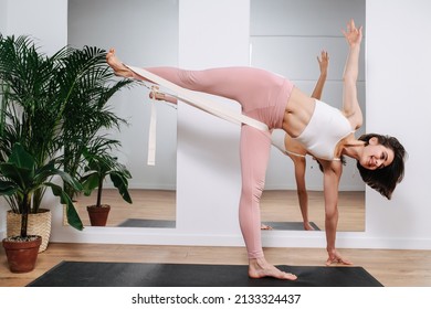 Cheerful Young Woman Stands On One Leg, Leaning Forward, Using Rubber Resistance Band. Yoga In A Living Room With Mirrors. She Is Wearing Pink Pants.