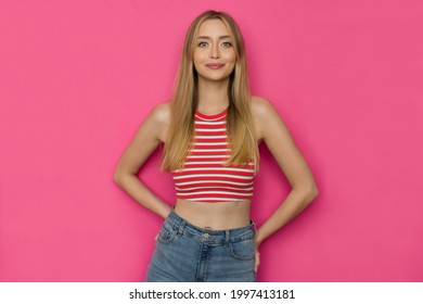 Cheerful young woman in striped tank top is posing with hands on hip. Front view. Waist up studio shot on pinkbackground.