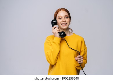 Cheerful Young Woman In Stylish Yellow Sweater Talking On Retro Phone And Looking At Camera Against Isolated White Background. Pretty Redhead Lady Model Emotionally Showing Facial Expressions.