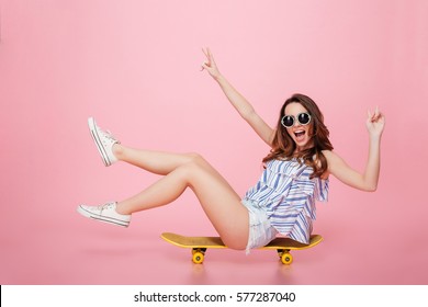 Cheerful young woman in sunglasses sitting on skateboard and having fun over pink background - Powered by Shutterstock