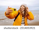 Cheerful young woman taking a selfie on a sandy beach with waves crashing in the background on a cloudy day. Selfie time. Cold windy weather. Outdoors. Travel concept.