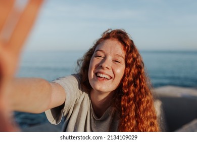 Cheerful young woman taking a selfie while standing next to the sea. Carefree young woman smiling happily at the camera. Outdoorsy young woman having fun in the summer sun. - Powered by Shutterstock