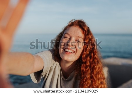 Cheerful young woman taking a selfie while standing next to the sea. Carefree young woman smiling happily at the camera. Outdoorsy young woman having fun in the summer sun.