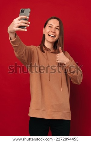 Cheerful young woman is taking a selfie with her phone while showing thumb up over red background.