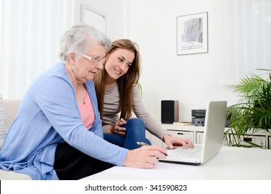 Cheerful Young Woman Teaching Computer To An Old Senior Woman At Home