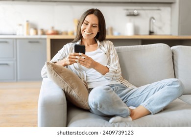 Cheerful young woman texting on her smartphone, smiling female sitting cross-legged on comfortable couch, happy millennial lady relaxing with mobile phone in cozy living room at home, copy space - Powered by Shutterstock