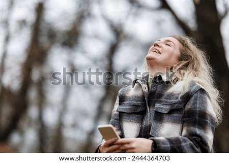 A cheerful young woman tilts her head back in a hearty laugh while holds a smartphone in her hands in an autumnal forest.