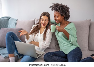 Cheerful Young Woman Using Laptop And Looking At Happy Friend While Sitting At Sofa And Discussing Homework Assignment In Cozy Room. Two Excited Women Shopping Online 