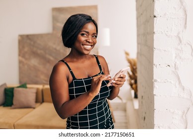 Cheerful young woman using smartphone. Smiling woman using app on cellphone at home. Beautiful girl relaxing while chatting on mobile phone and looking at camera.