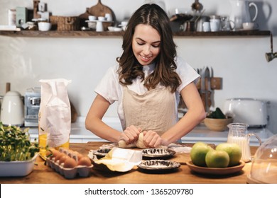 Cheerful young woman wearing apron preparing dough for an apple pie at the kitchen at home - Powered by Shutterstock