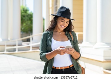 Cheerful Young Woman Wearing Black Hat In City Street Typing A Message. Hispanic Trendy Girl Browsing Internet On Phone Outdoor. Happy Stylish Girl Using Smartphone While Travelling The City.