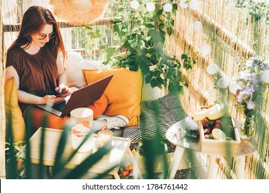 Cheerful young woman wearing brown blouse and sunglasses working on modern laptop sitting on the balcony with lemonade on sunny day. 
