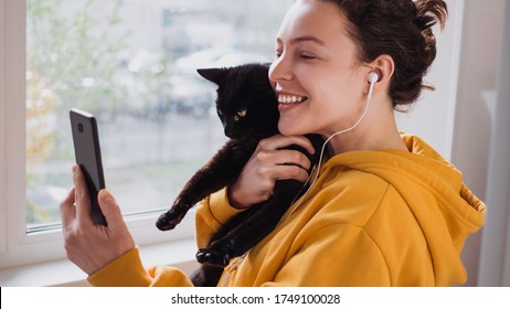 Cheerful Young Woman Wearing Headphones Holds Black Pet Cat Using Smartphone For Video Call, Gesturing Hi To Friend Or Parent.Caucasian Girl In Yellow Hoodie Making Selfie,sharing Data On Social 