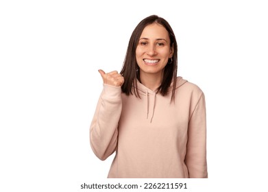 Cheerful young woman wearing a light pink hoodie points aside with her thumb over white background. - Powered by Shutterstock