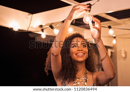 Cheerful young woman wearing party dress changing bulb light in patio. Happy smiling girl making preparation of party by adding lights outdoor. Happy beautiful woman enjoying fixing bulbs in backyard.