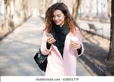 Cheerful young woman wearing pink coat using her phone in the sunny city street and drinking take away coffee in paper cup.