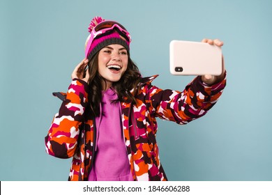 Cheerful Young Woman Wearing Snowboard Gear Taking A Selfie Isolated Over Blue Backgorund