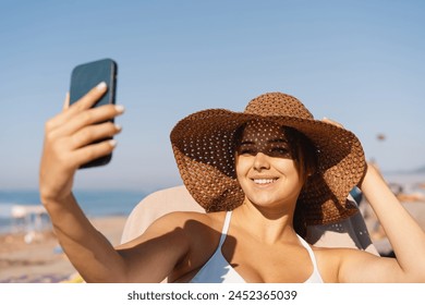 A cheerful young woman wearing a wide-brimmed straw hat and a white bikini takes a selfie with her smartphone, enjoying the bright sunshine by the ocean shore with a clear blue sky overhead. - Powered by Shutterstock