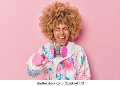 Cheerful Young Woman Wears Radiation Protective Suit Gas Mask Around Neck Points Index Finger At Camera And Laughs Positively Isolated Over Pink Background. Female Worker At Nuclear Power Plant