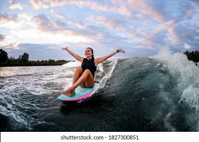 cheerful young young woman with wet hair while riding wave while sitting on surf style wakeboard with hands up