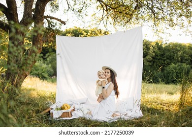 Cheerful Young Woman In White Summer Dress And Hat Lifting Up Six Month Old Baby Girl While Playing Sitting On Blanket Outdoors. Mother With Daughter Enjoying Time Spending Together On Fresh Air.