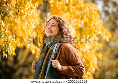 Cheerful young woman with winter scarf relaxing at park with yellow trees in background. Smiling beautiful girl enjoying warm sunny weather in autumn season. Happy pretty natural woman laughing.